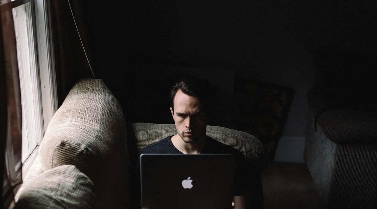 A person working on a laptop from a sofa beside a bright window